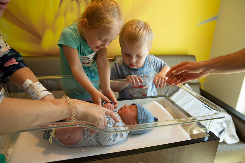 a newborn baby in a hospital cradle, while his siblings look at him over the edge