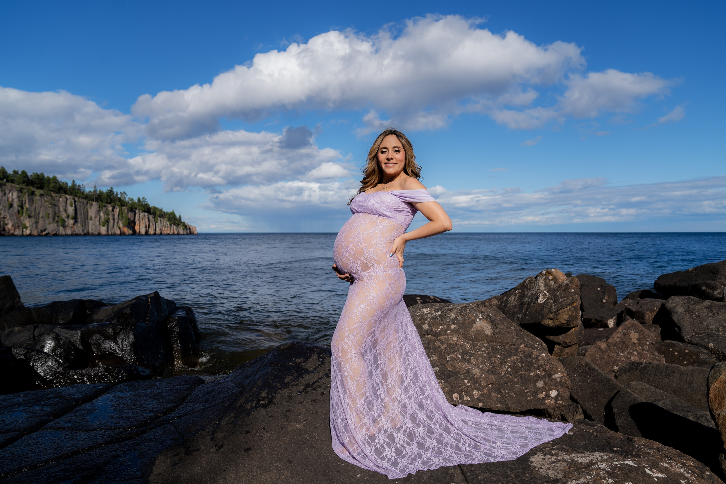 Pregnant woman in a translucent lilac gown standing in front of the sea