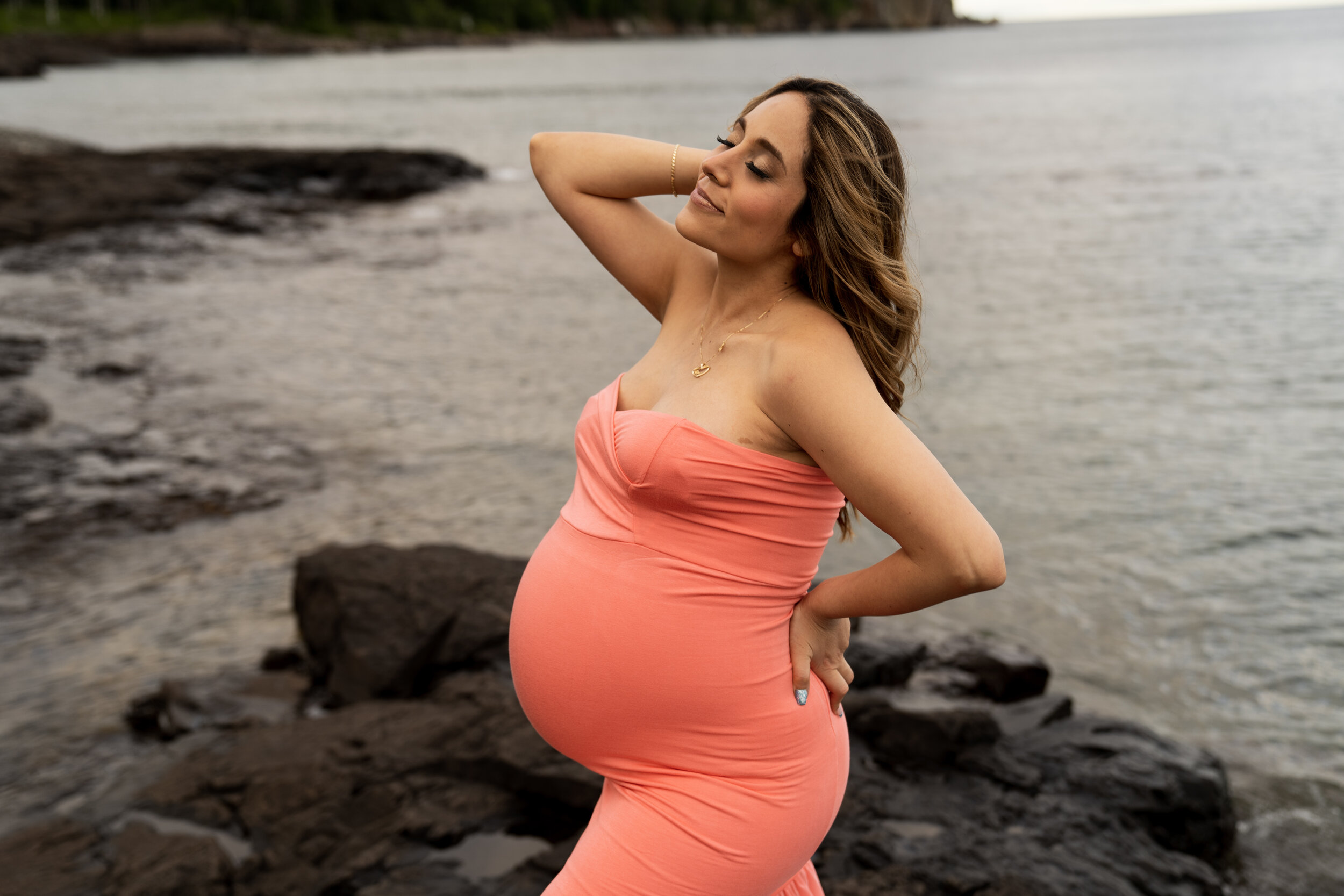 Pregnant woman in a coral dress standing in front of the ocean on a rocky outcropping