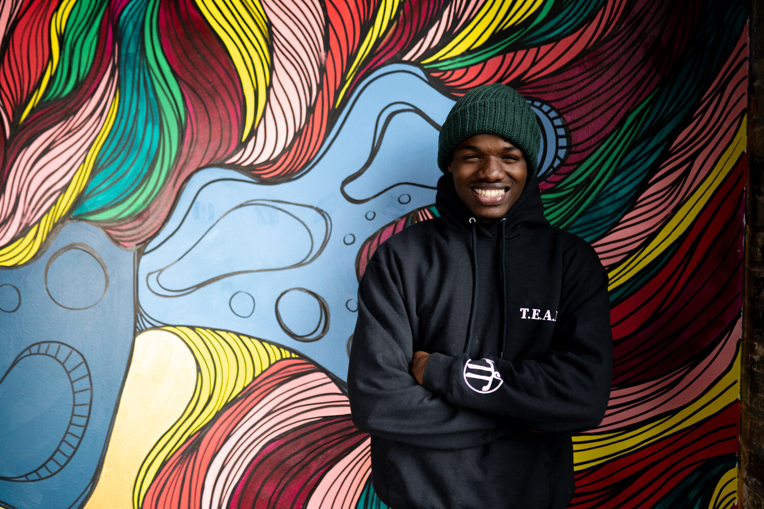 Man in all black smiling with folded arms in front of a bright flowing mural