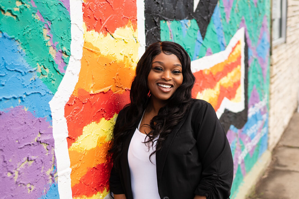 Black woman smiling widely wearing a professional blazer, leaning against a colorful mural in Minneapolis MN