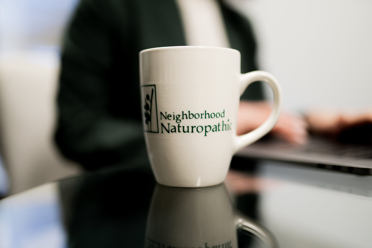 White mug placed on a table, the mug reads "Neighborhood Naturopathic" in forest green lettering