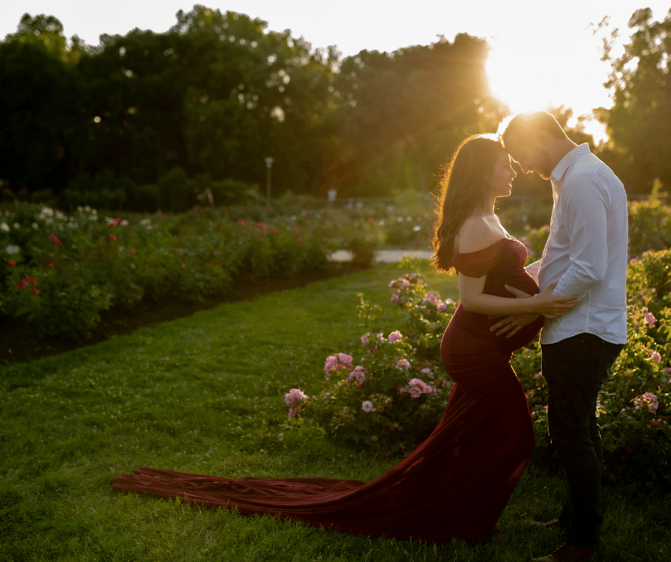 father-to-be holding his partner's belly. She's wearing an elegant red dress and the sun is setting behind them