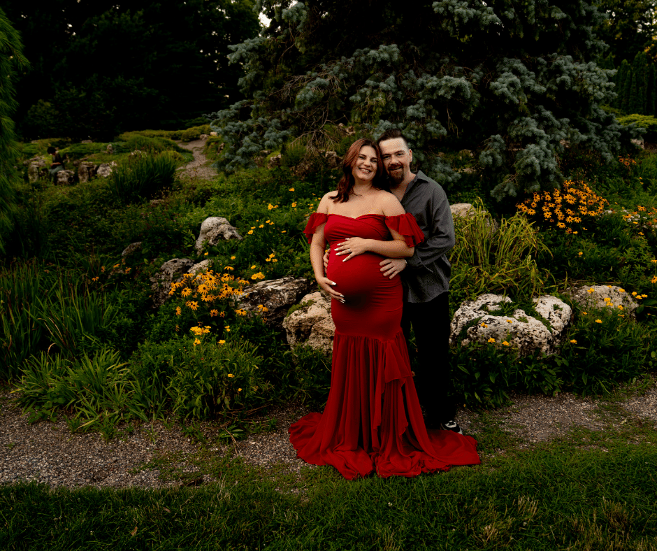 Mother-to-be wearing an elegant red dress posed with her husband at Lyndale rose garden minneapolis mn