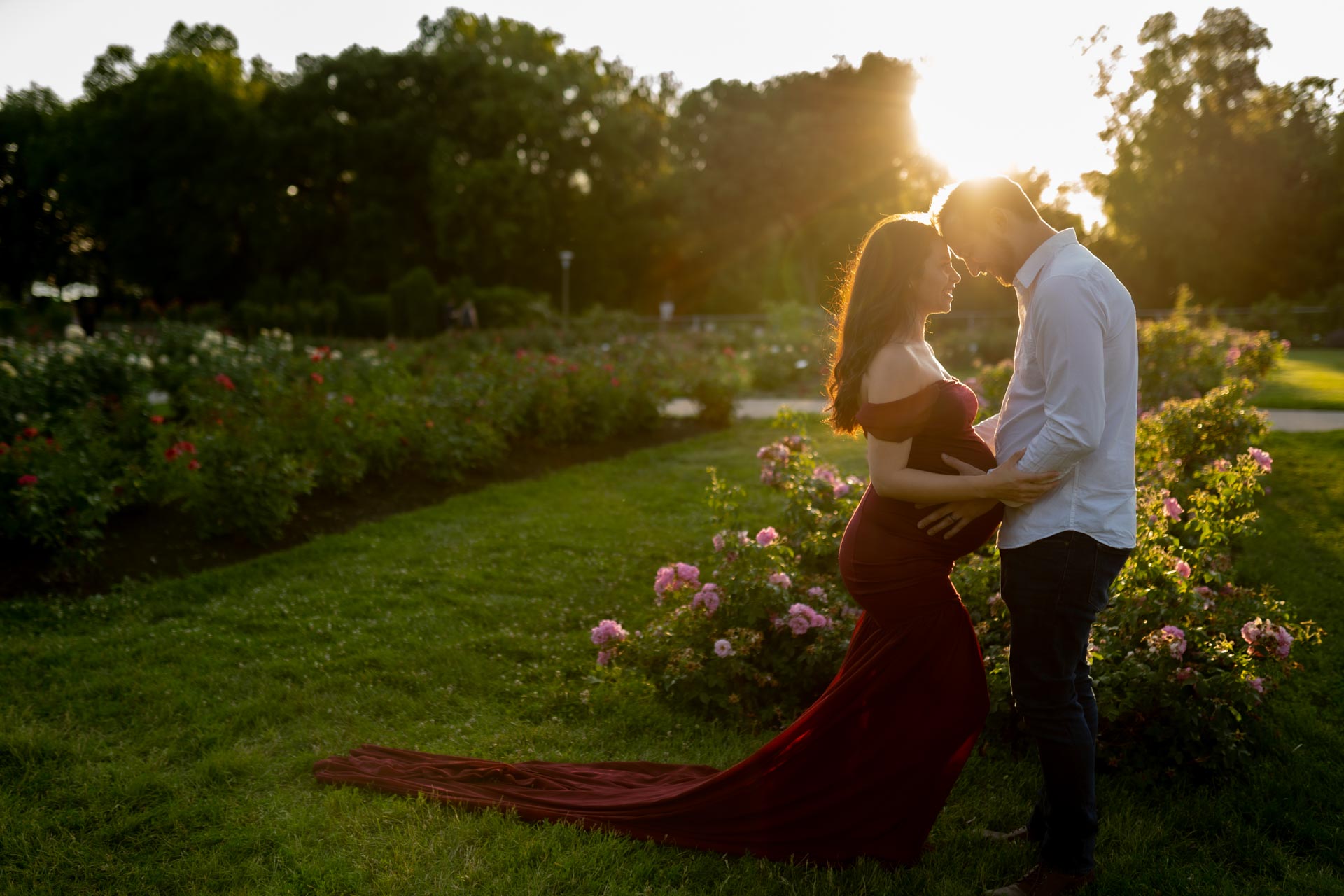 father-to-be holding his partner's belly. She's wearing an elegant red dress and the sun is setting behind them