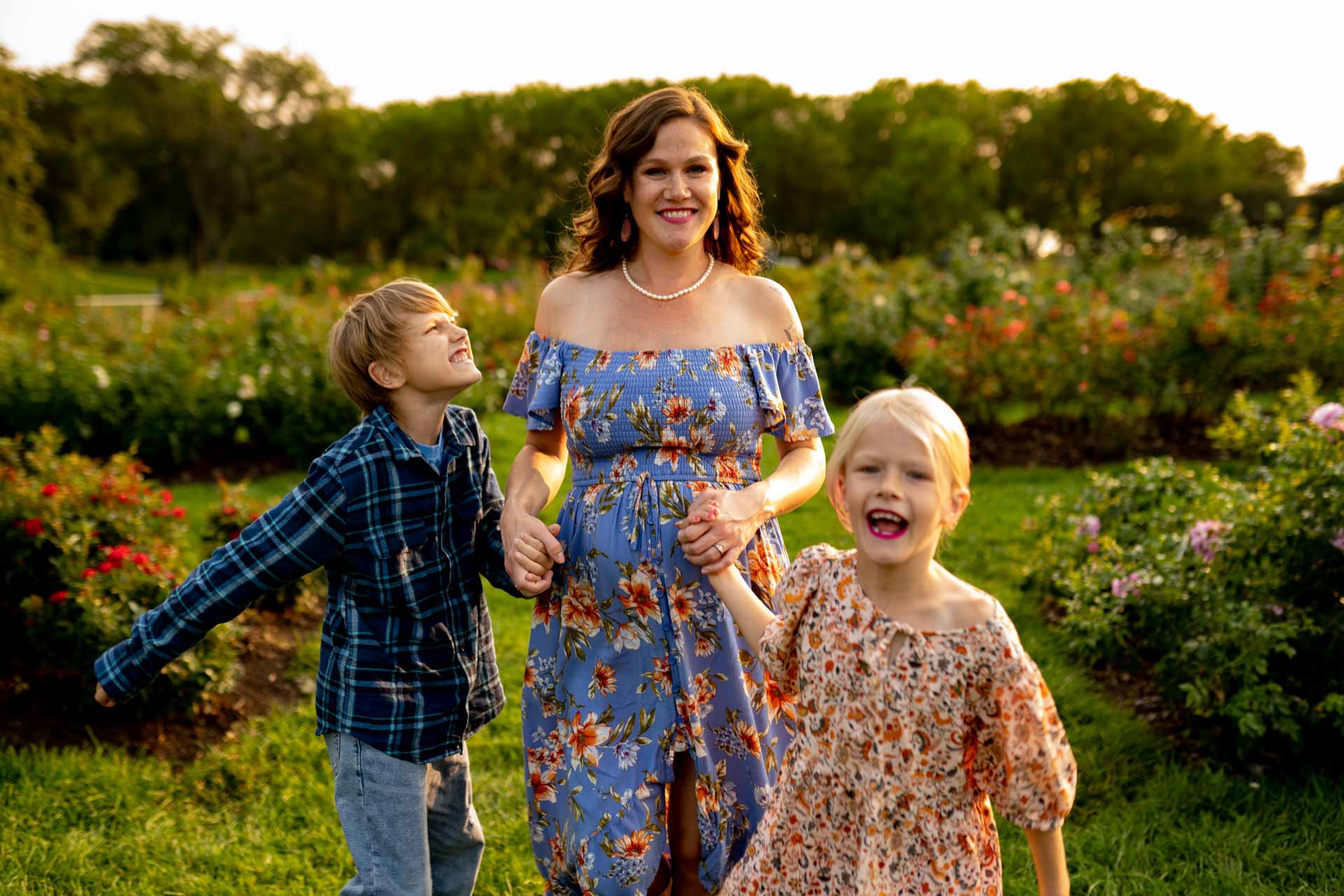 Mother in a floral dress holding her children's hands while walking through lush greenery