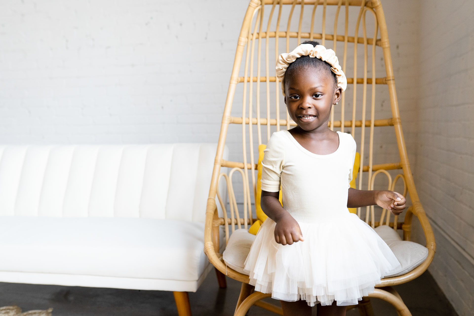 a little girl posed in front of a wicker chair, wearing all white