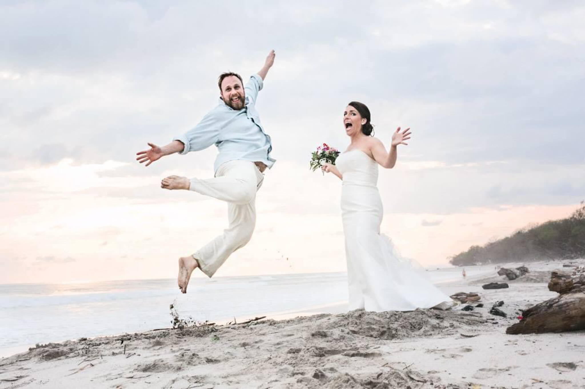 Jessica Strobel in her wedding dress on a beach, while her husband jumps into the air wearing linen