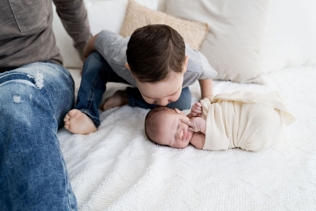 A toddler boy in blue jeans kisses the head of his newborn sibling while they sleep on a bed in a white swaddle