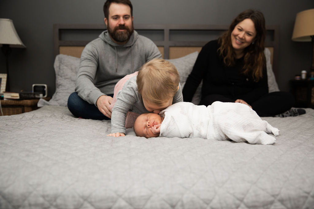 A toddler boy in a grey shirt leans over and kisses his newborn sibling who is sleeping on a bed with mom and dad sitting behind them on the bed minneapolis doulas