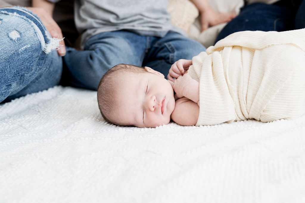 A newborn baby sleeps in a white swaddle on a bed with it's family behind looking on wearing blue jeans minneapolis doulas