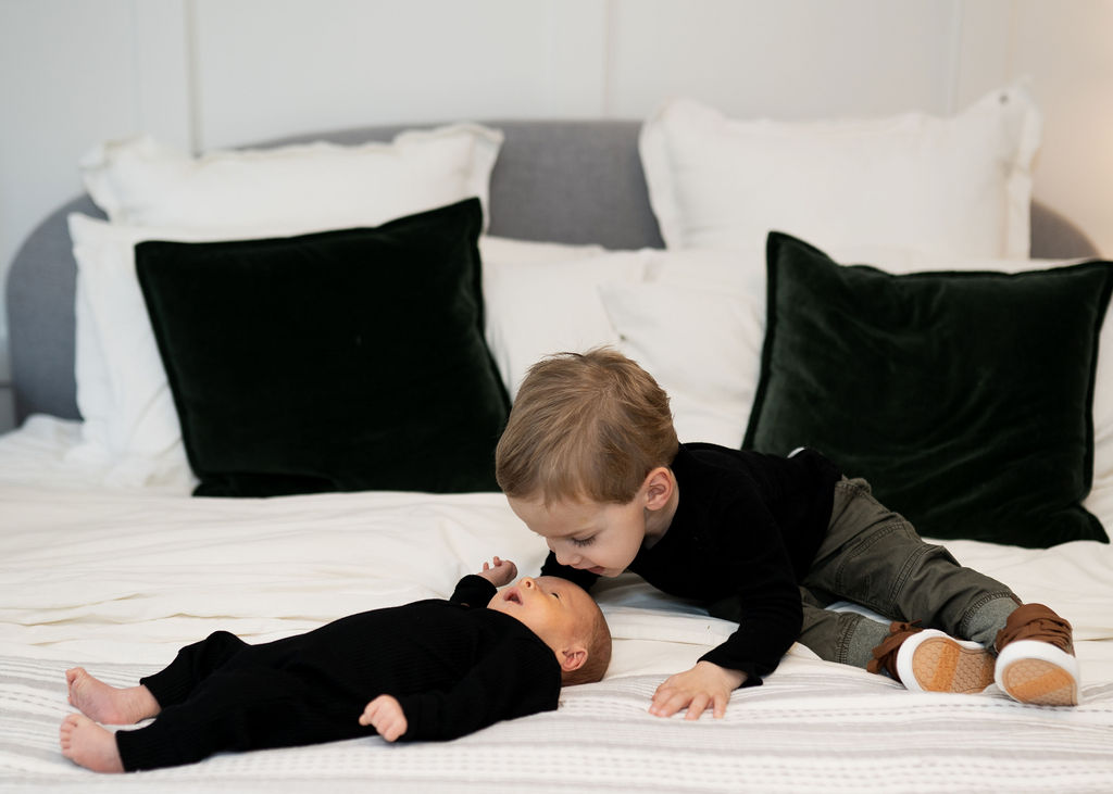 A toddler boy sits on a bed in a black shirt and plays with his newborn sibling wearing a black onesie laying next to him twin cities birth center
