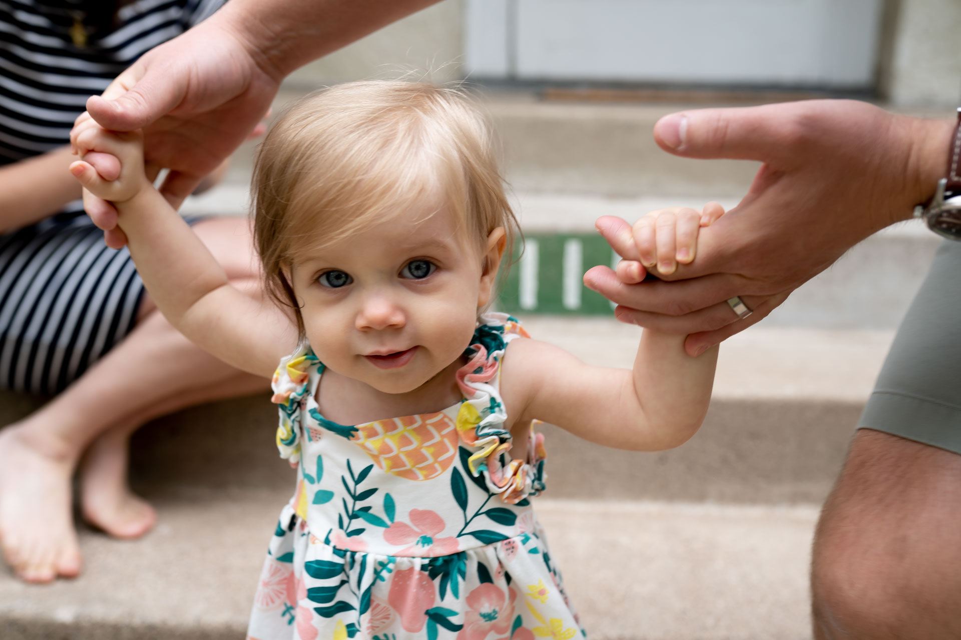 toddler in bright flower dress holding adults hands looking at the camera smiling