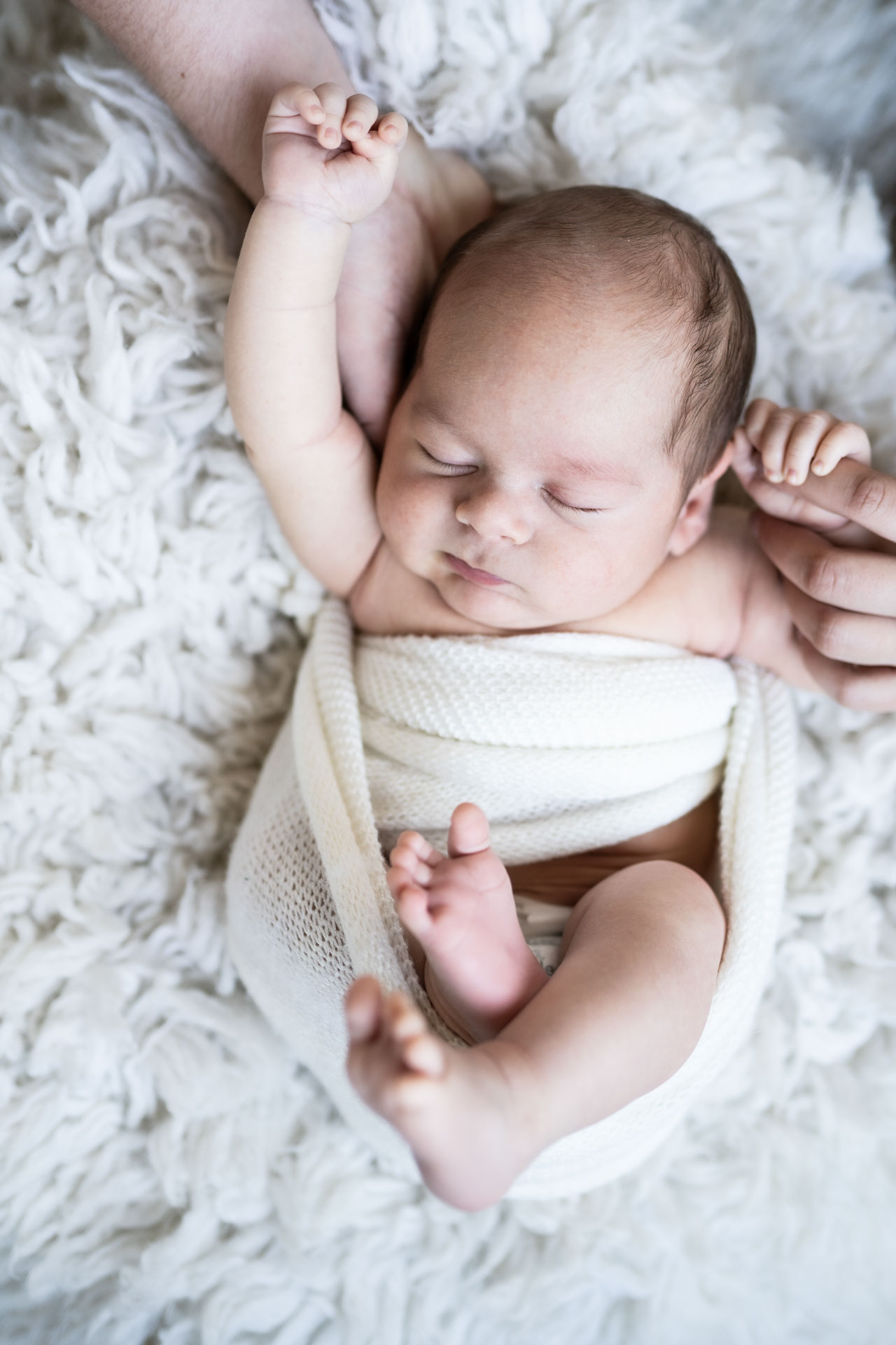 newborn baby boy on white rug wearing white wrap holding moms hand