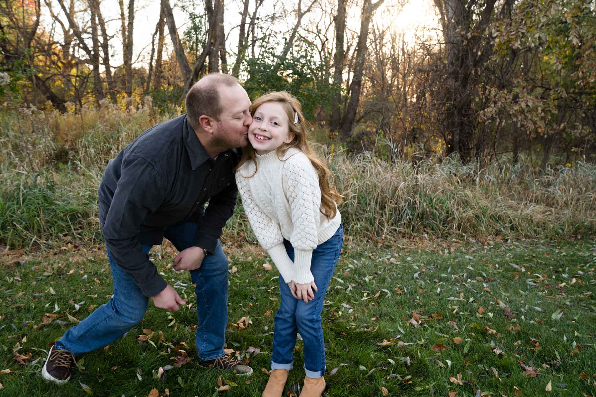 dad and daughter wearing jeans and sweaters during a fall photo shoot. dad giving daughter a kiss on the cheek