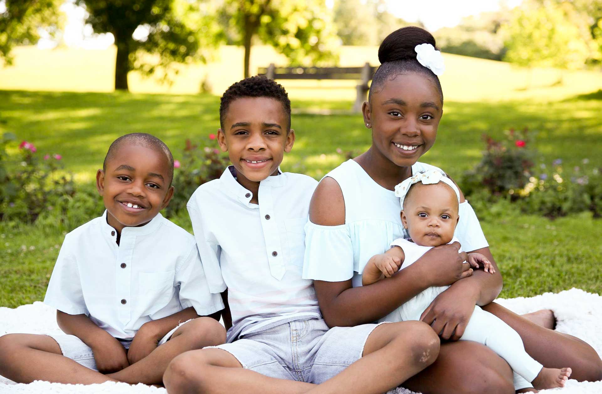 four African American siblings wearing all white, seated in a lush green Minneapolis park, smiling
