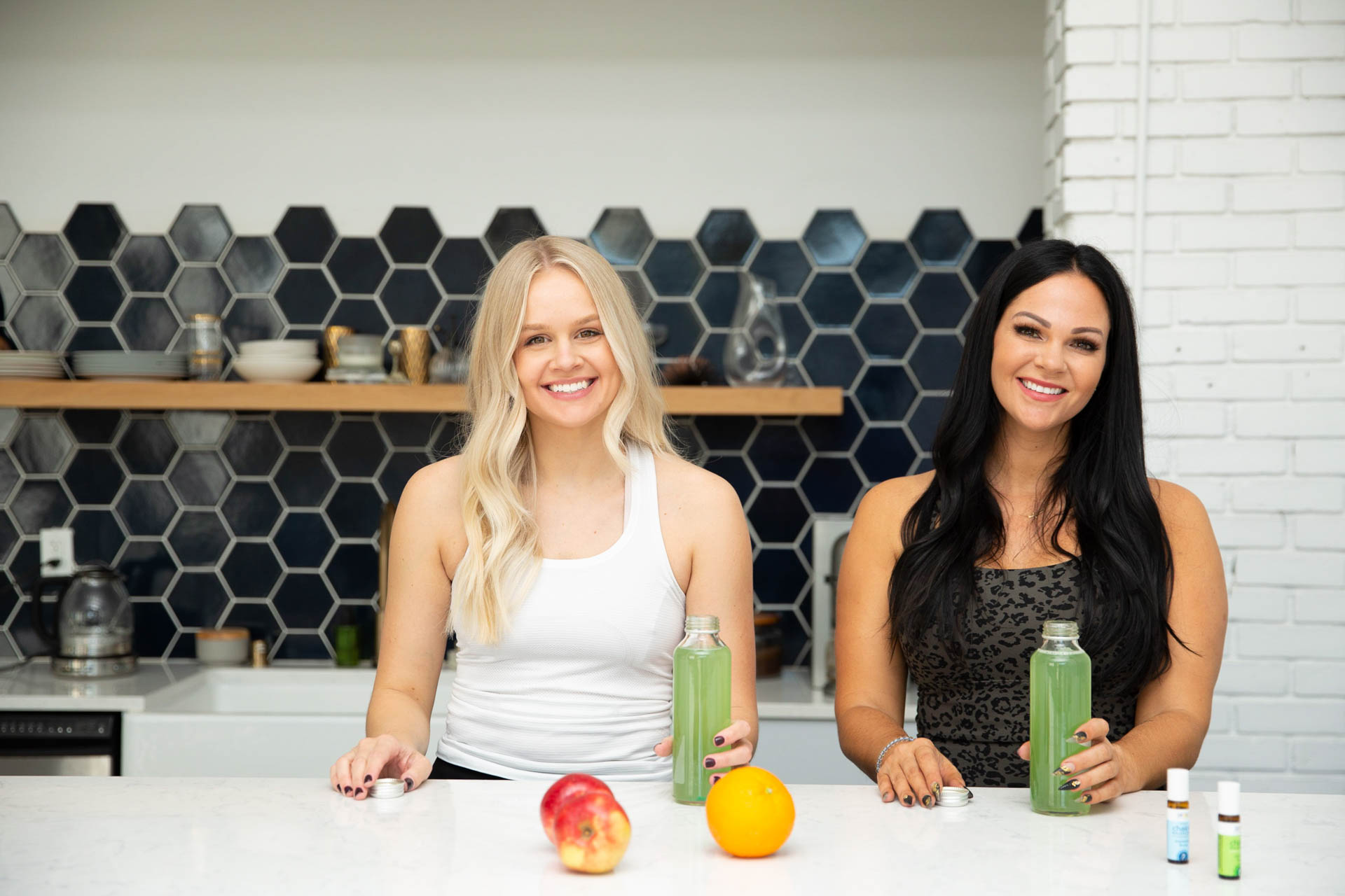 two women standing and smiling at the kitchen counter holding green smoothie bottles