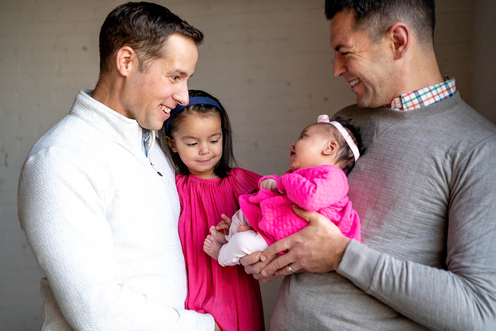 Two dads in neutral outfits holding their yound daughters in hot pink dresses, and smiling