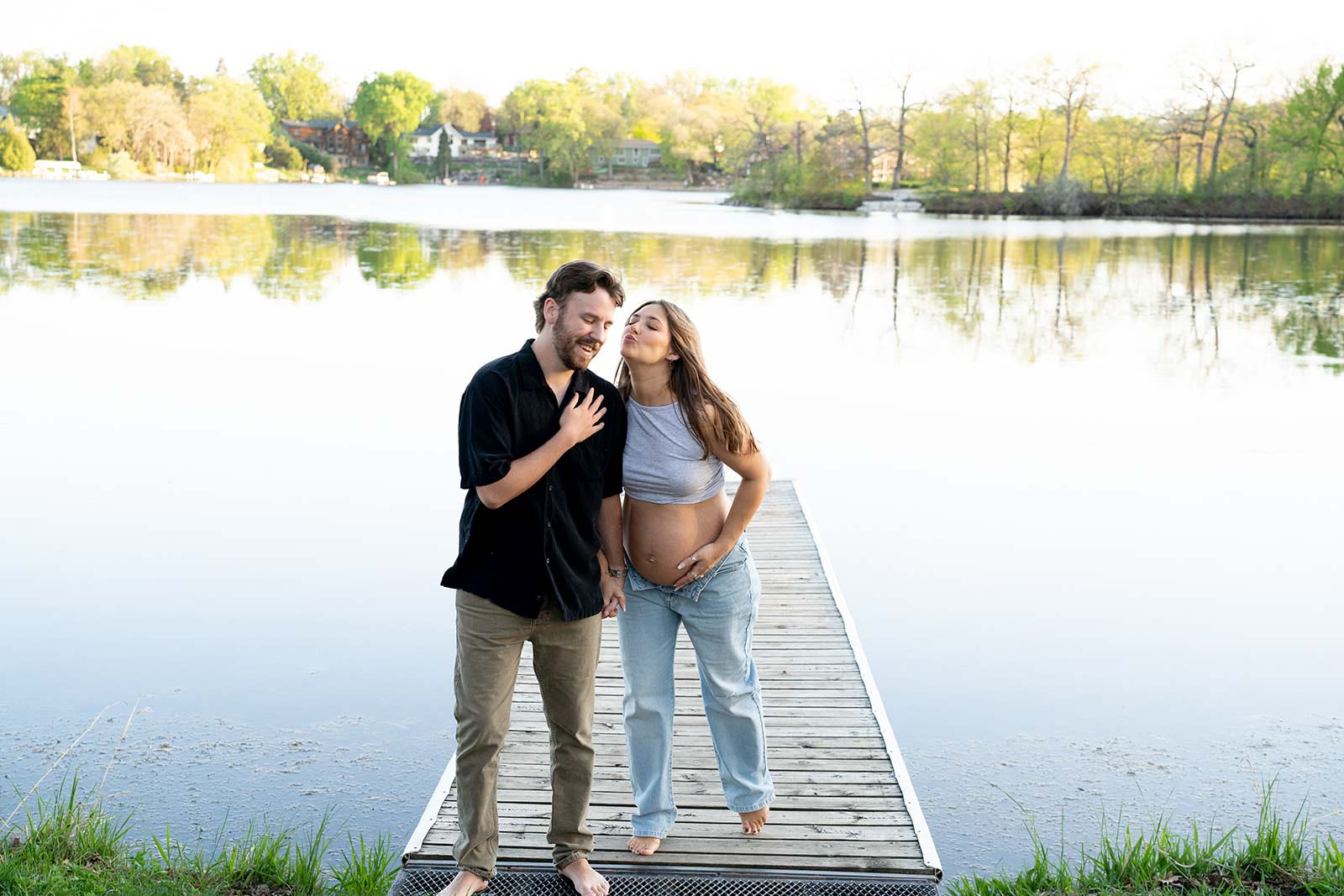 a man and woman in casual clothing, goofing around on a dock over a lake, with her pregnant belly exposed through her denim jeans