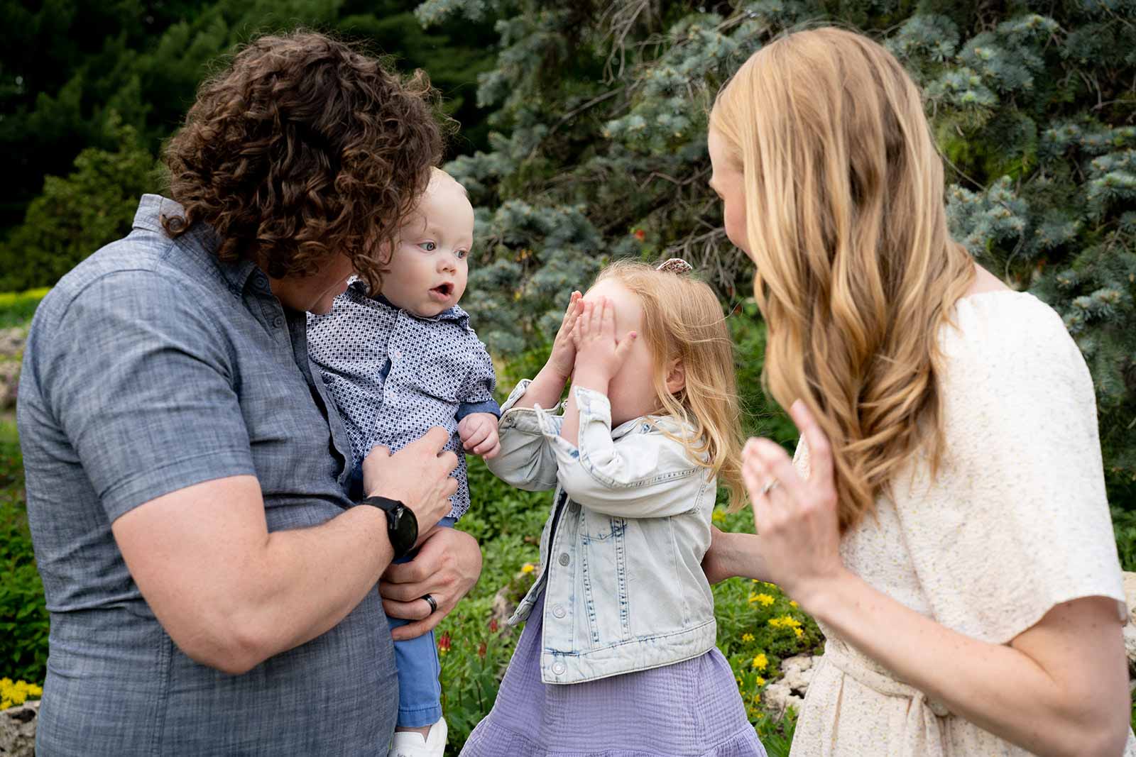 a young girl playing peekaboo with her younger brother, while both are being held by their mom and dad