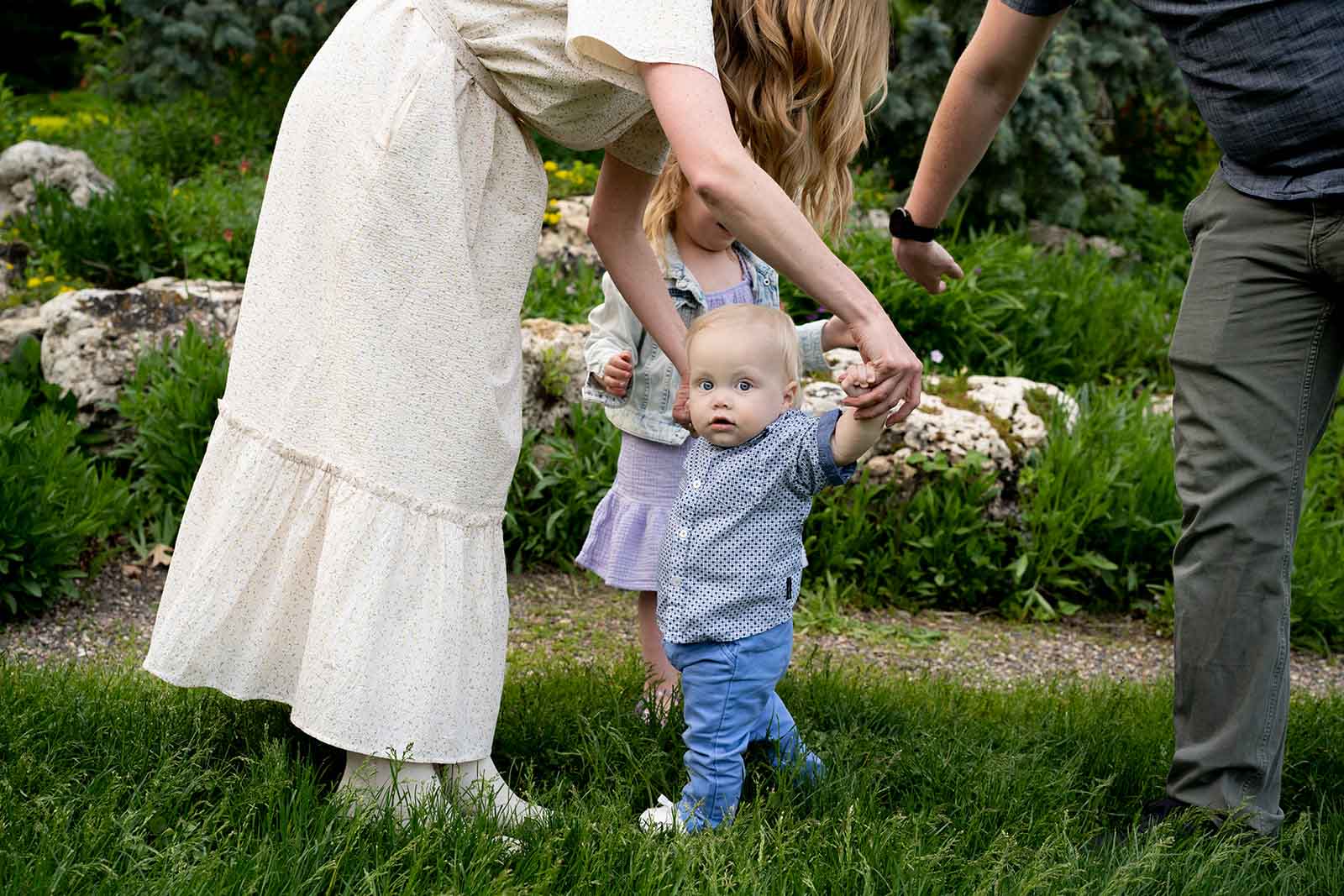 Linden A toddler in a casual button down, having his arms held up to help him walk, by his mother who is wearing a cream dress
