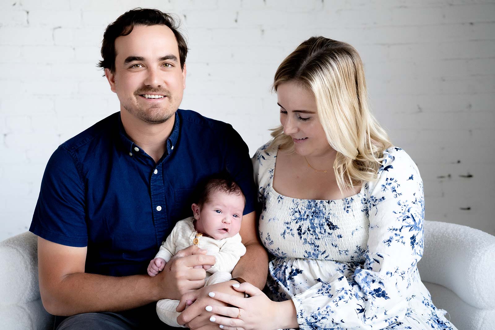 A father looking into the camera and whiling, with his newborn baby on his knee, while the mother looks emotively down at the baby