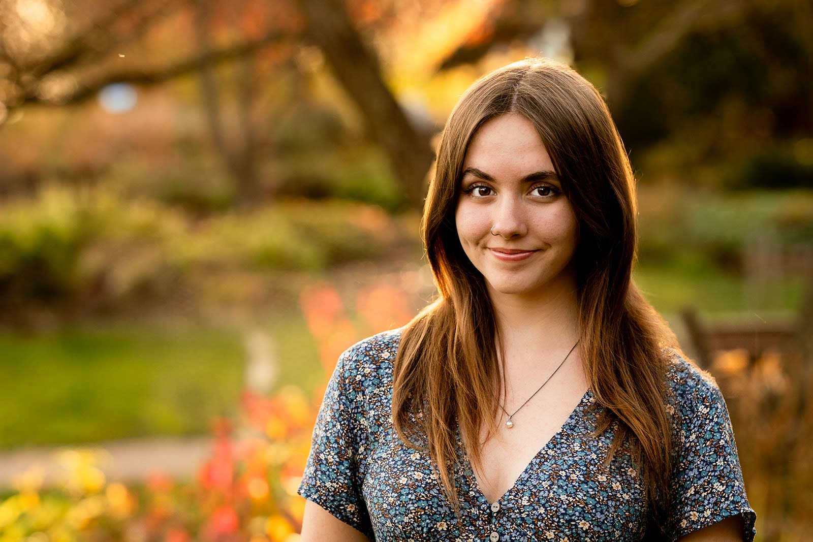 High school senior in a floral dress smiling for the camera in front of a golden hour autumn outdoor scene