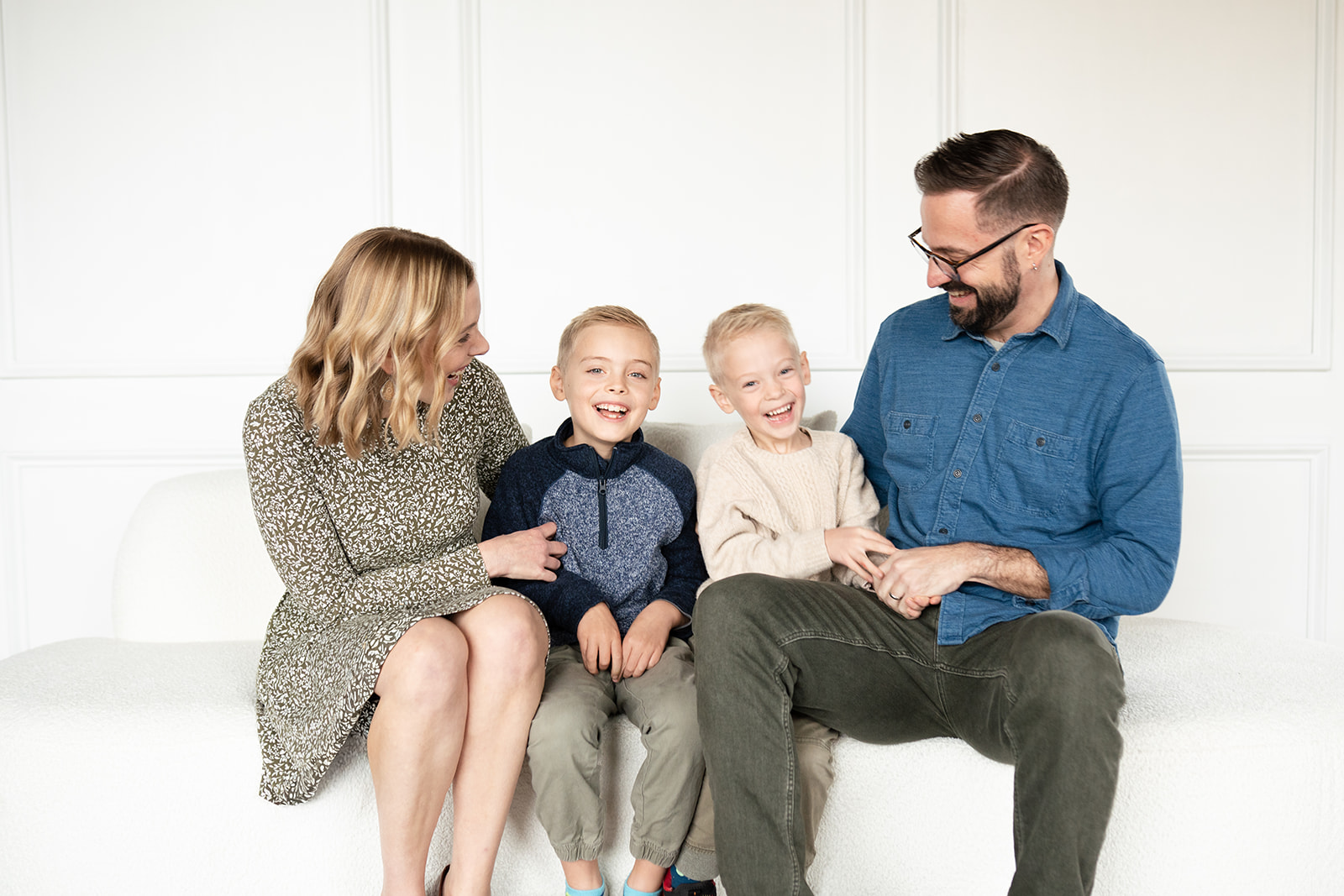family of four sitting on white couch with white background laughing. Mom and dad are looking at the children while the young boys are looking at the camera