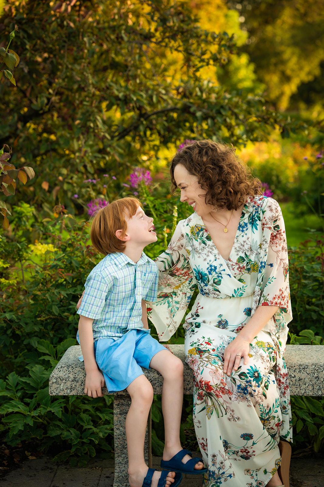 mother is floral dress looking as son dressed in button down and blue shirt. Seated on cement bench surround by beautiful greenery.