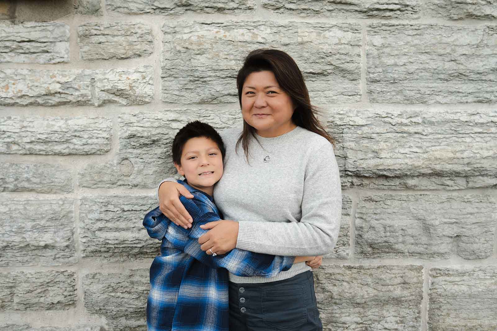 Mother and son posing in front of a grey stone wall, embracing and smiling