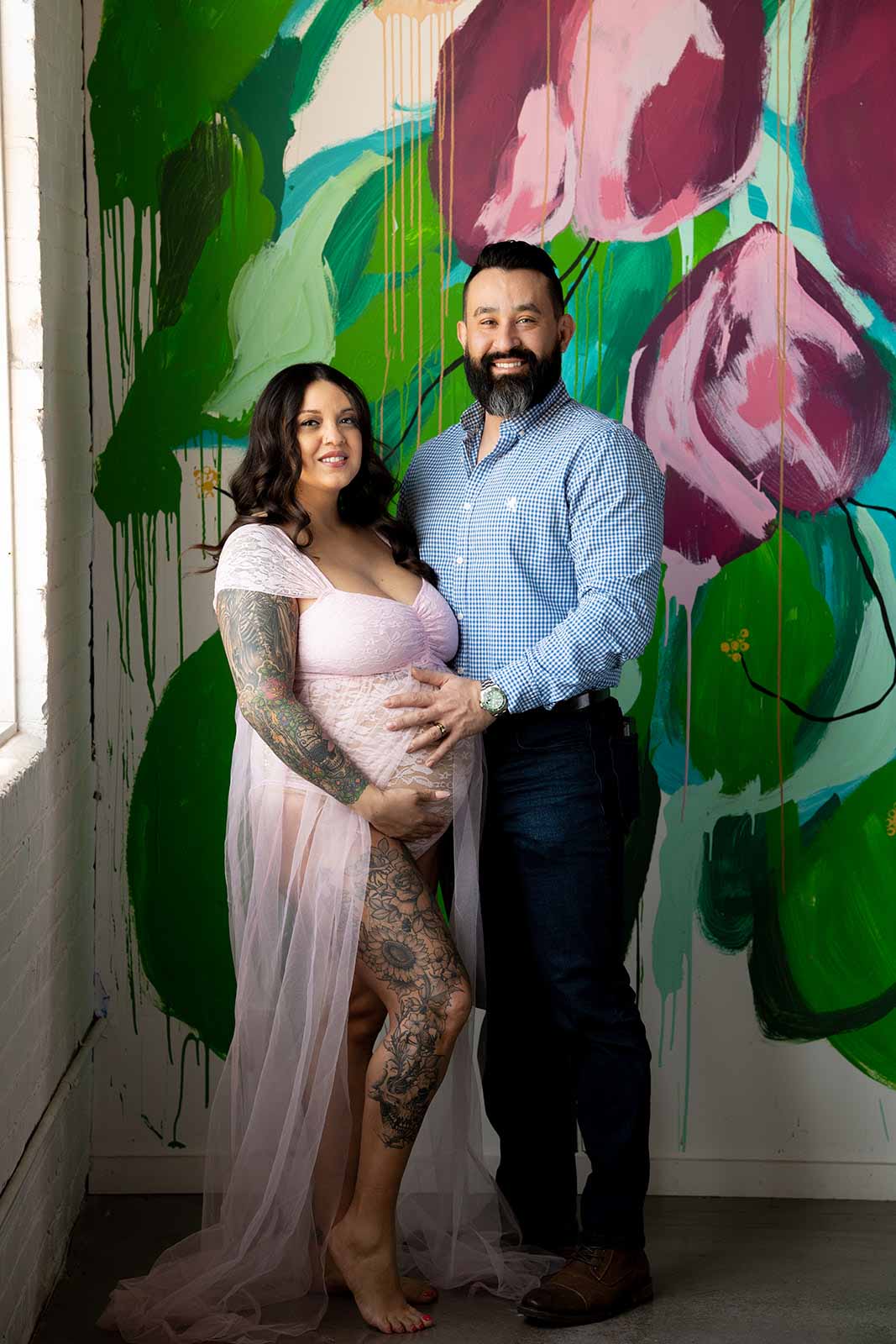a pregnant woman with her husband standing behind her and holding her belly, looking seriously and emotionally out of the window both wearing black